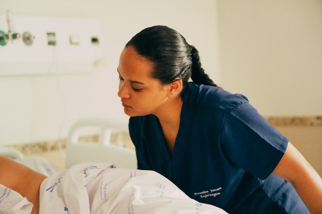 A nurse in dark blue scrubs leans over a patient in a hospital bed, attending to them during a clinical placement.