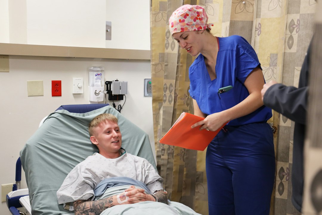 A nurse in blue scrubs holds a red clinical folder while speaking with a patient in a hospital bed