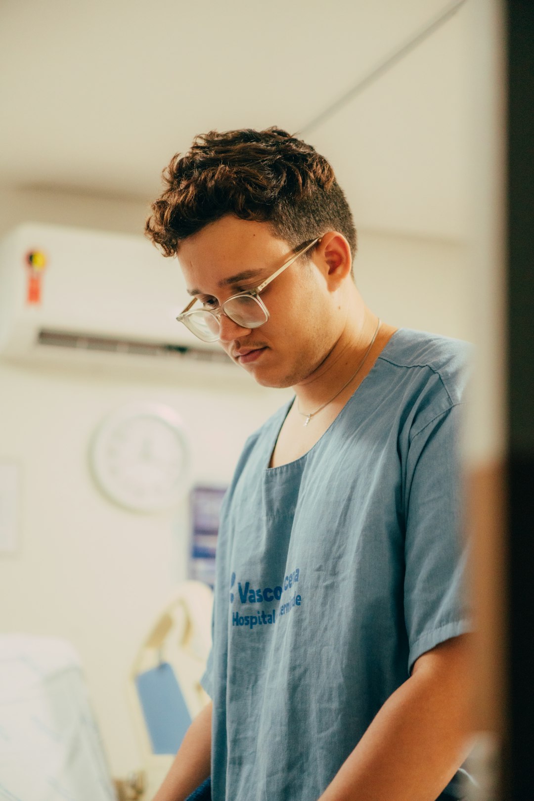 A young nursing student in light blue scrubs stands in a clinical training setting, focused on the task.