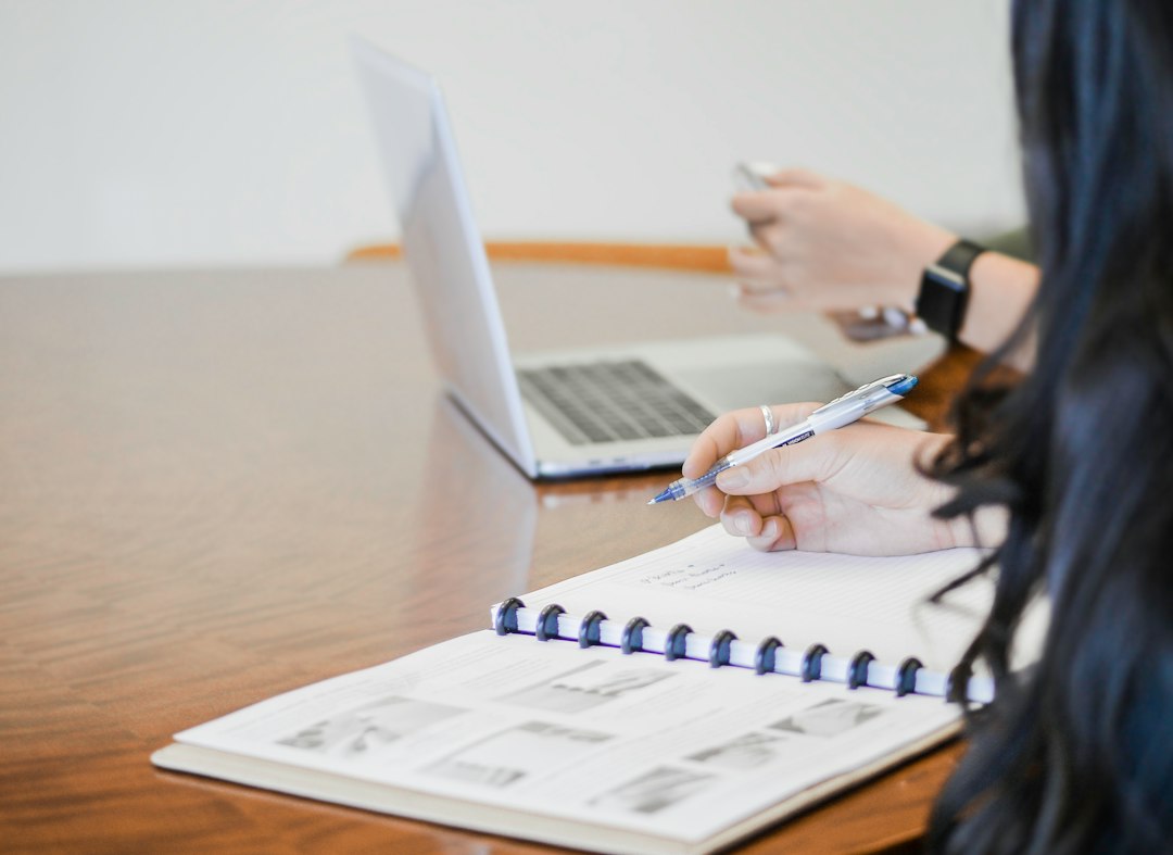 Coordinator writing notes in a spiral notebook beside an open laptop, illustrating how good evidence collection is documented and tracked